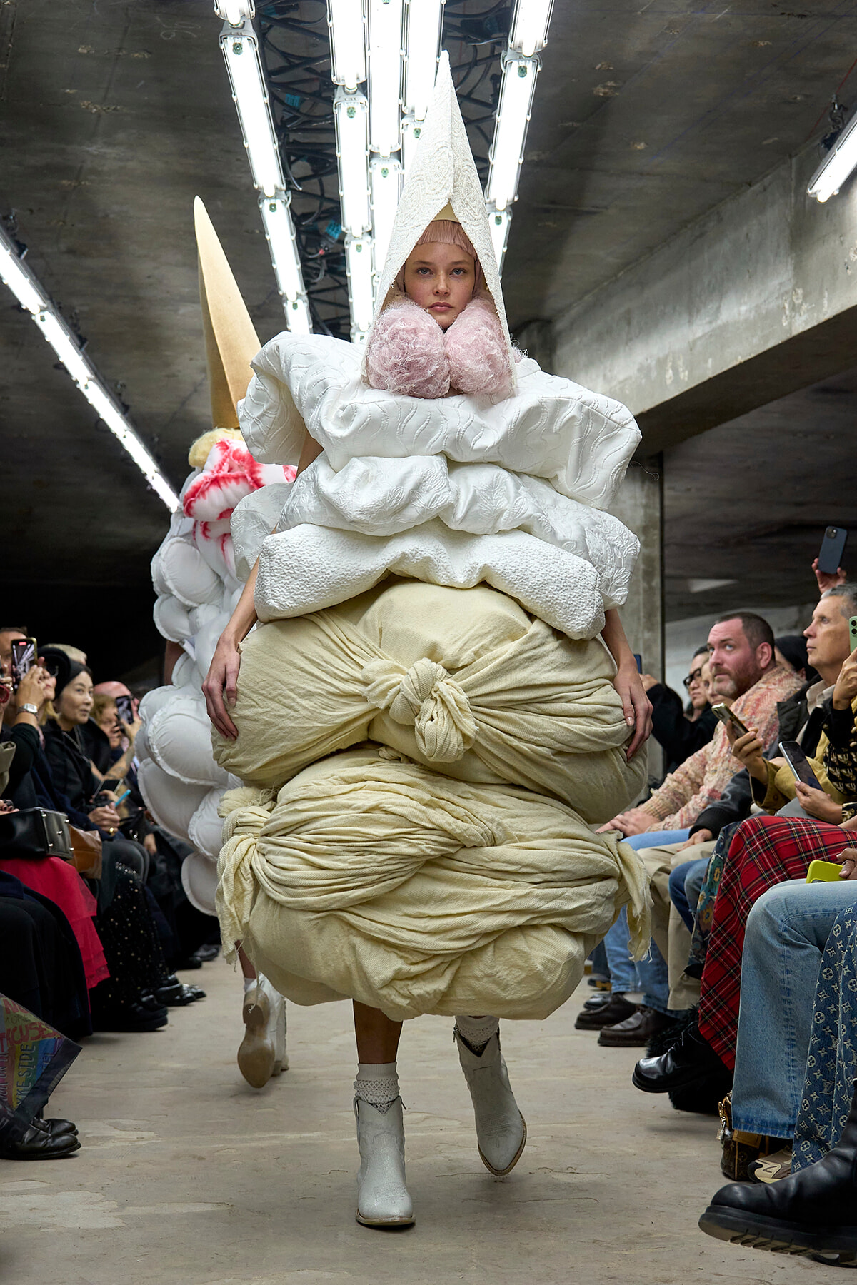 Model walks the runway in an oversized sculptural outfit: white quilted top with large padded layers and a cream knotted skirt, wearing a tall cone headpiece and pink fluffy collar, industrial venue runway with spectators taking photos.