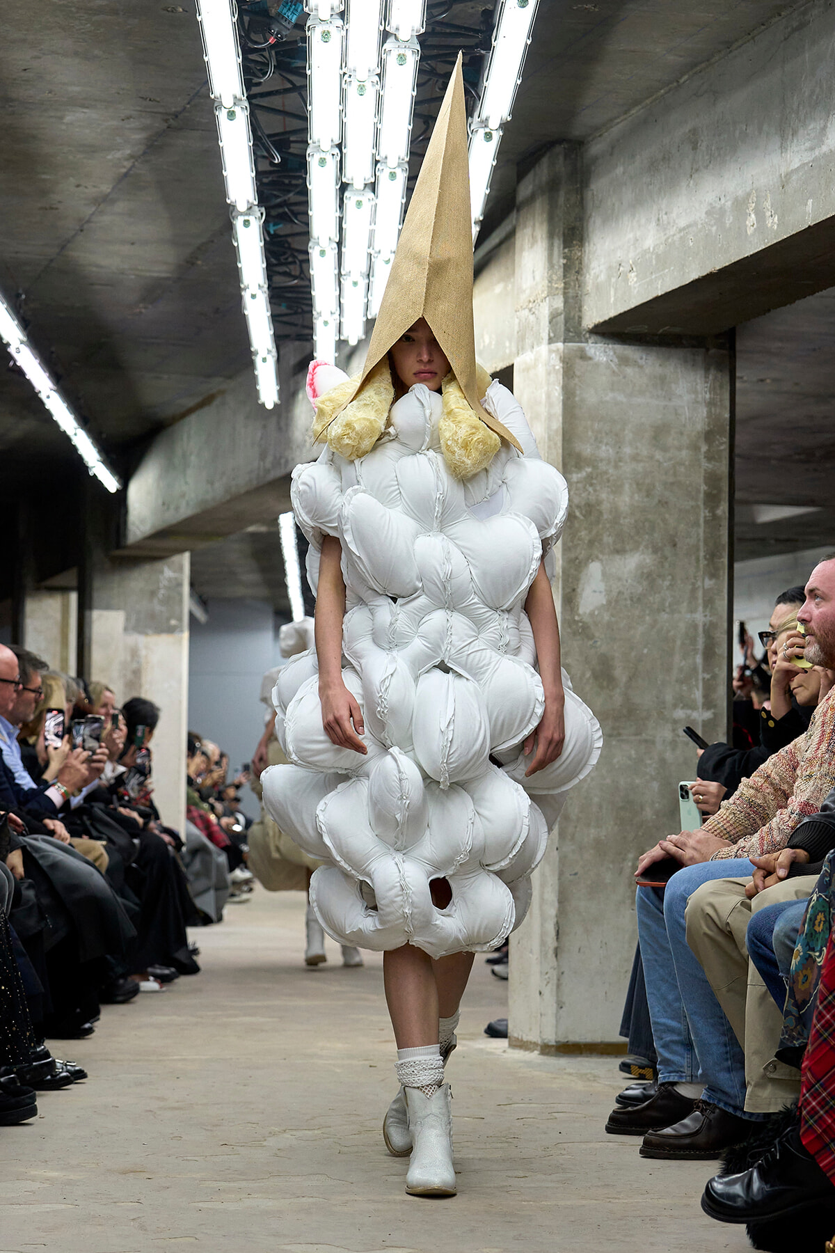 Model walks a runway in a voluminous white, cloud-like dress with a tall beige cone hat in an industrial venue, as seated audience watches.