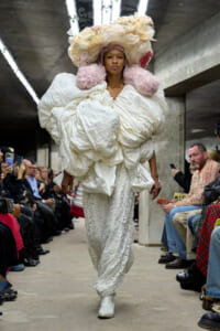 Model walking a runway in an oversized white sculptural coat with pink puffed shoulder accents and a dramatic headpiece.