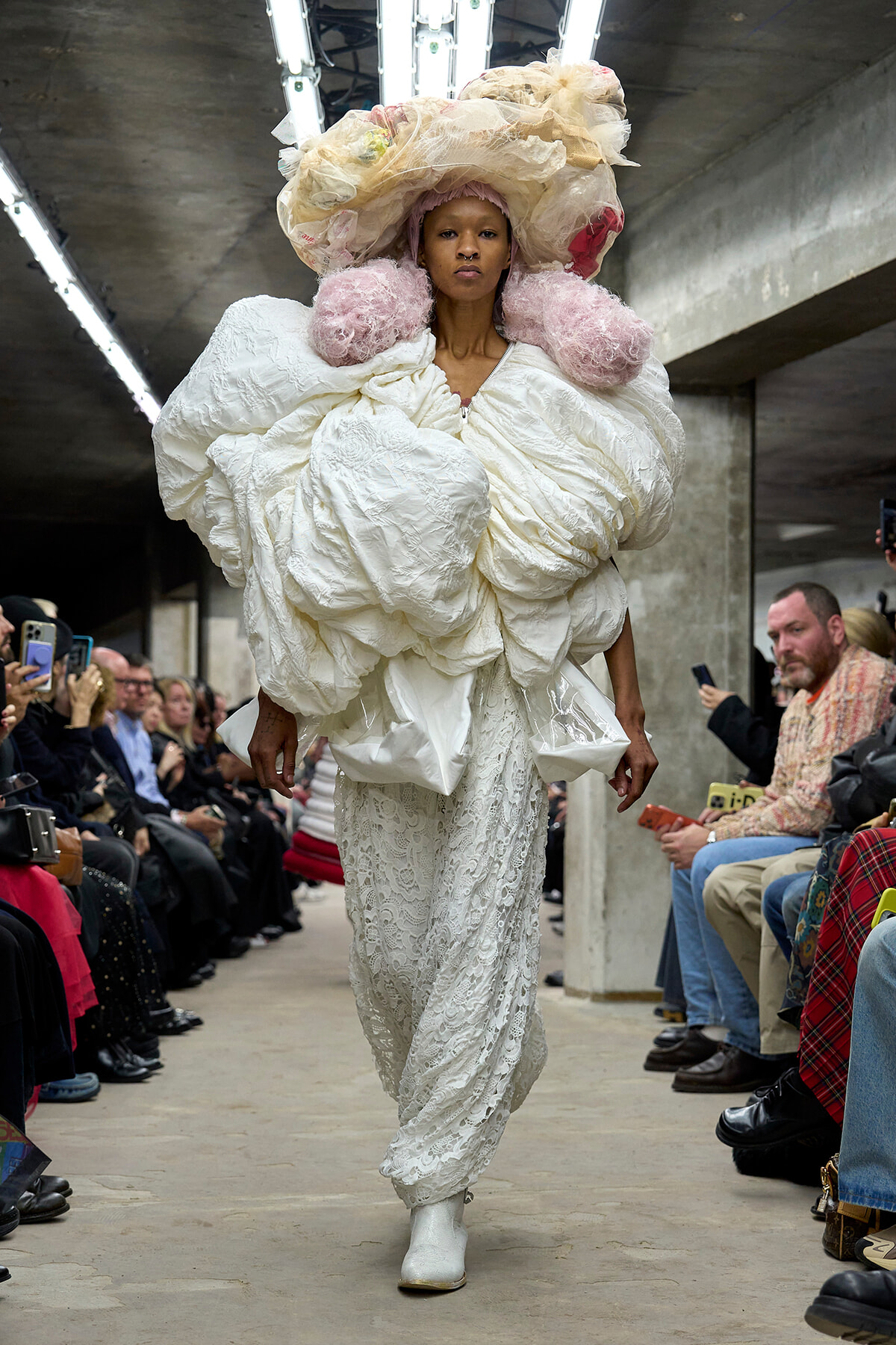 Model walking a runway in an oversized white sculptural coat with pink puffed shoulder accents and a dramatic headpiece.