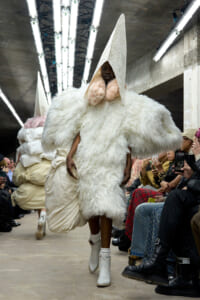 Model on a runway wearing a voluminous white fluffy outfit with a tall cone hood; audience on both sides watching and filming the look.