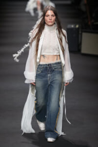 Model on a fashion runway wearing a sheer white long coat, a cropped lace blouse, and faded denim jeans with a relaxed fit.