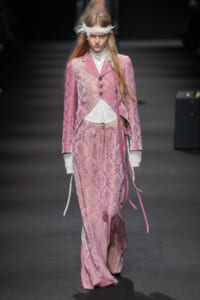 Model on a runway wearing a pink lace brocade suit with a white top and feather headband for a high-fashion show.