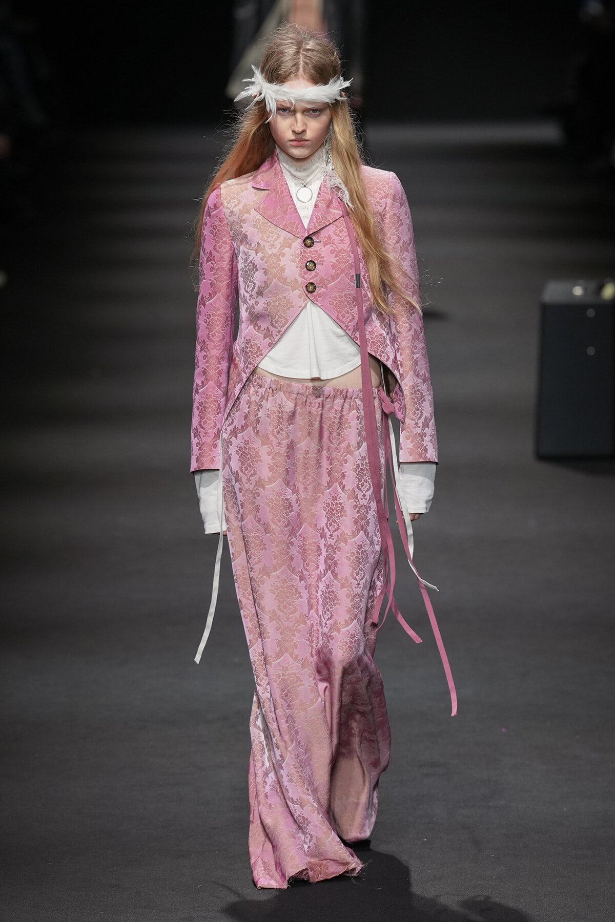 Model on a runway wearing a pink lace brocade suit with a white top and feather headband for a high-fashion show.