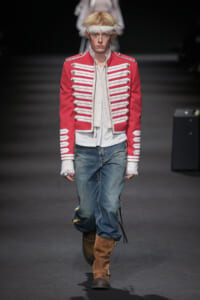 Male runway model wearing a red military-style jacket with white braiding, white shirt, loose blue jeans, and brown knee-high boots on a dark runway backdrop.