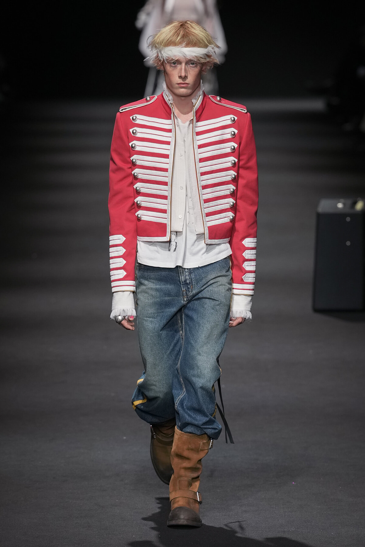 Male runway model wearing a red military-style jacket with white braiding, white shirt, loose blue jeans, and brown knee-high boots on a dark runway backdrop.