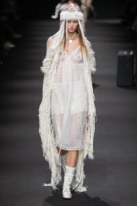 Model on a runway wearing a sheer white lace dress with a long fringed feathered cloak and matching headpiece.