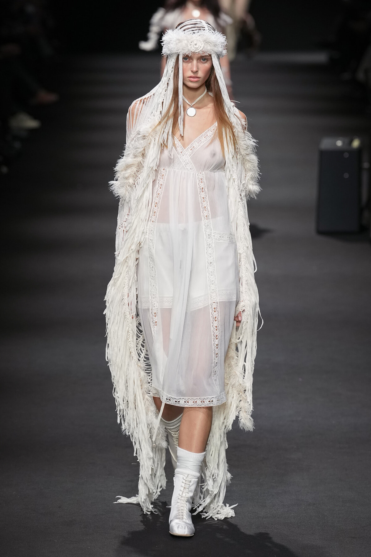 Model on a runway wearing a sheer white lace dress with a long fringed feathered cloak and matching headpiece.