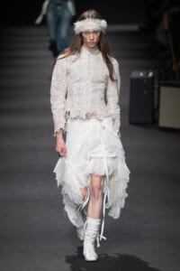 Model on a runway wearing an ivory crochet blouse with scalloped hem and a voluminous layered white skirt, white boots, and a fluffy feather headband.