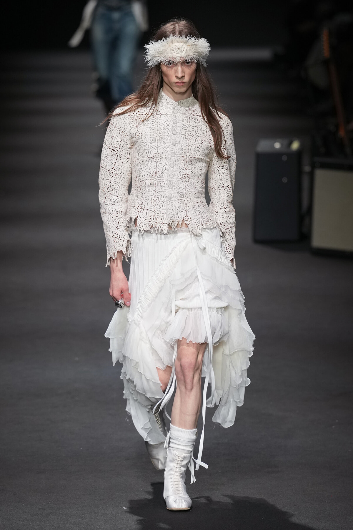 Model on a runway wearing an ivory crochet blouse with scalloped hem and a voluminous layered white skirt, white boots, and a fluffy feather headband.