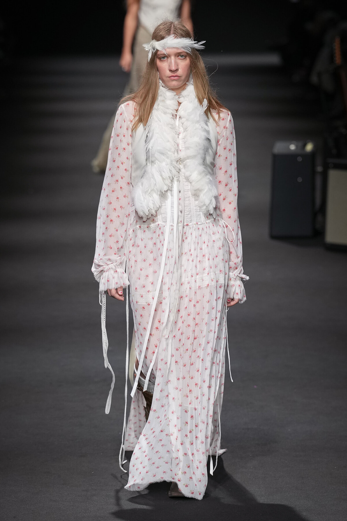 Model on a runway wearing a long white floral dress with a white feather scarf and headband.