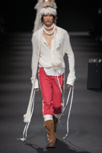 Male model walks the runway in a loose white shirt, red sporty pants with white piping, brown boots, and fluffy knit accessories.