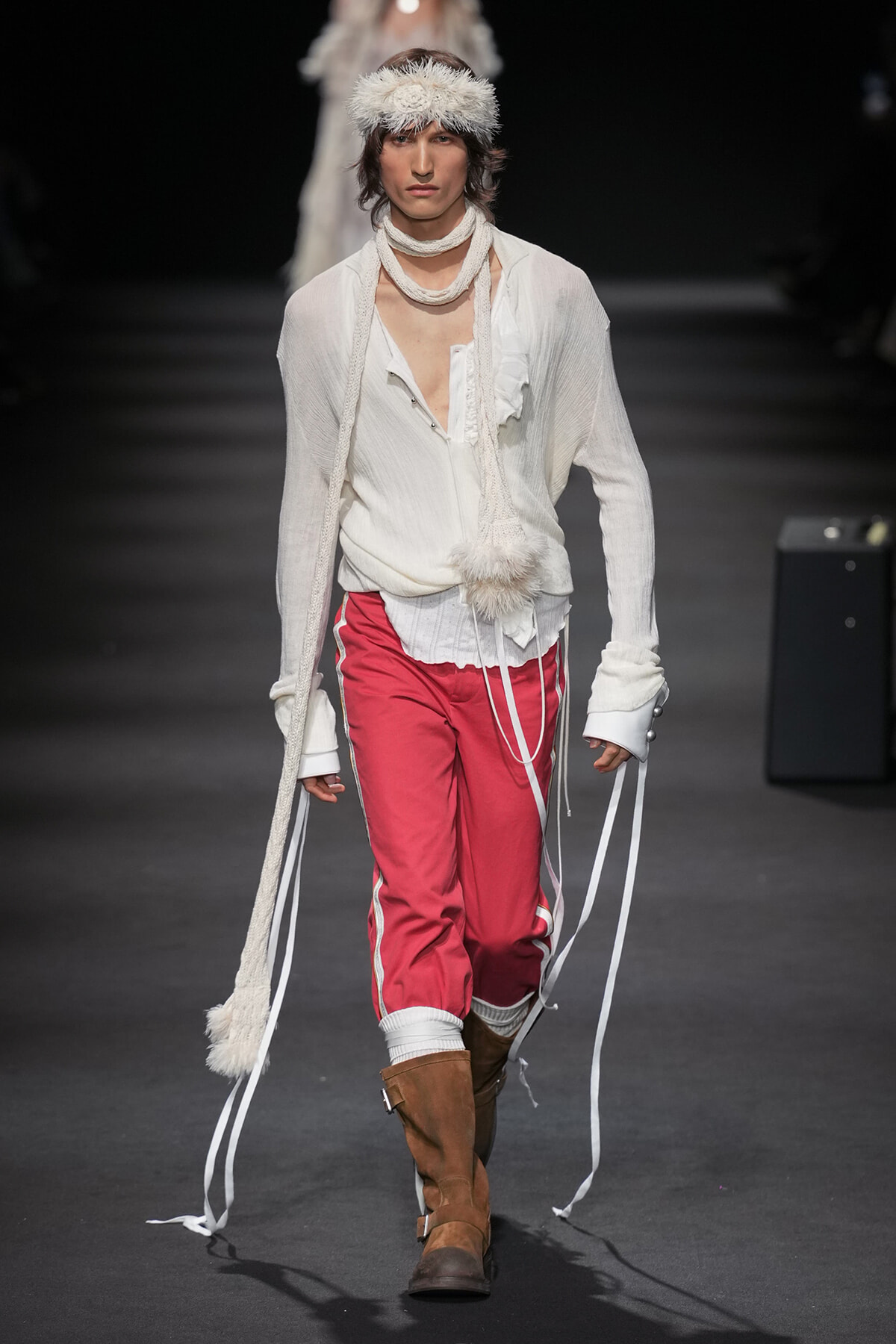 Male model walks the runway in a loose white shirt, red sporty pants with white piping, brown boots, and fluffy knit accessories.