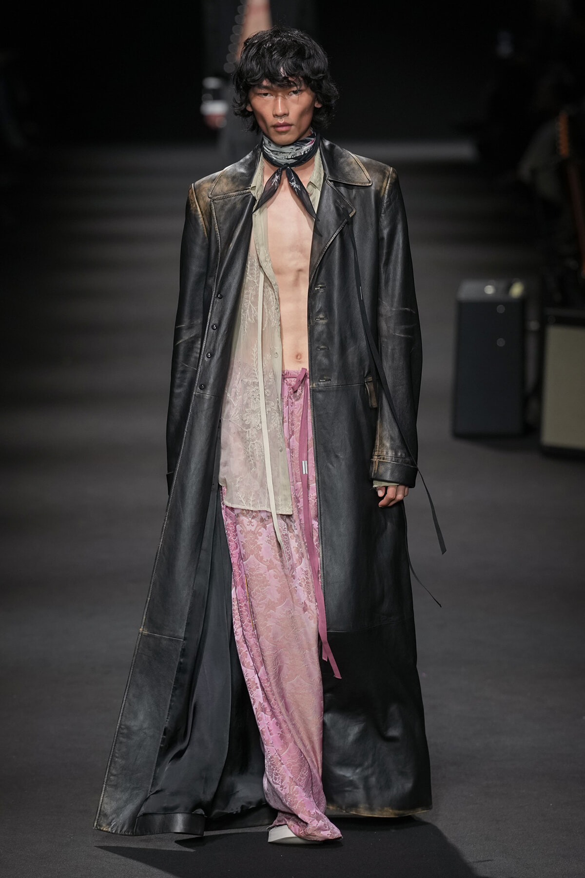 Male model on a runway wearing a long black leather coat over a sheer beige shirt and pink lace-patterned pants, with a neck scarf.