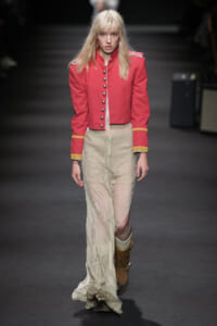 Model walking the runway in a red cropped military-style jacket with gold trim and metal buttons, paired with a sheer cream lace skirt and brown boots.