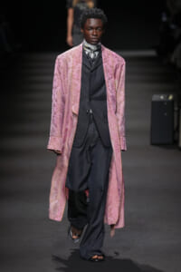 Male model on a runway wearing a long pink brocade coat over a dark pinstripe suit and scarf, stepping forward confidently.