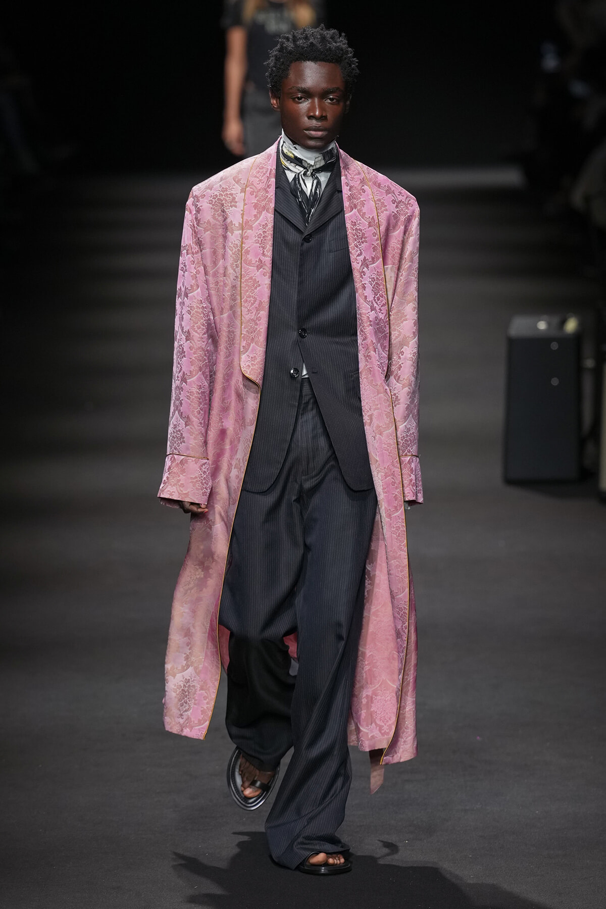 Male model on a runway wearing a long pink brocade coat over a dark pinstripe suit and scarf, stepping forward confidently.