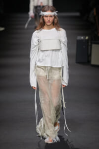 Model on the runway wearing a white long-sleeve top with a corset-style bustier over it and sheer lace pants, accessorized with a feather headband.
