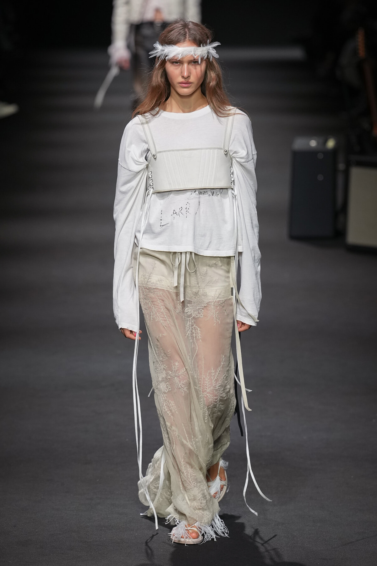 Model on the runway wearing a white long-sleeve top with a corset-style bustier over it and sheer lace pants, accessorized with a feather headband.