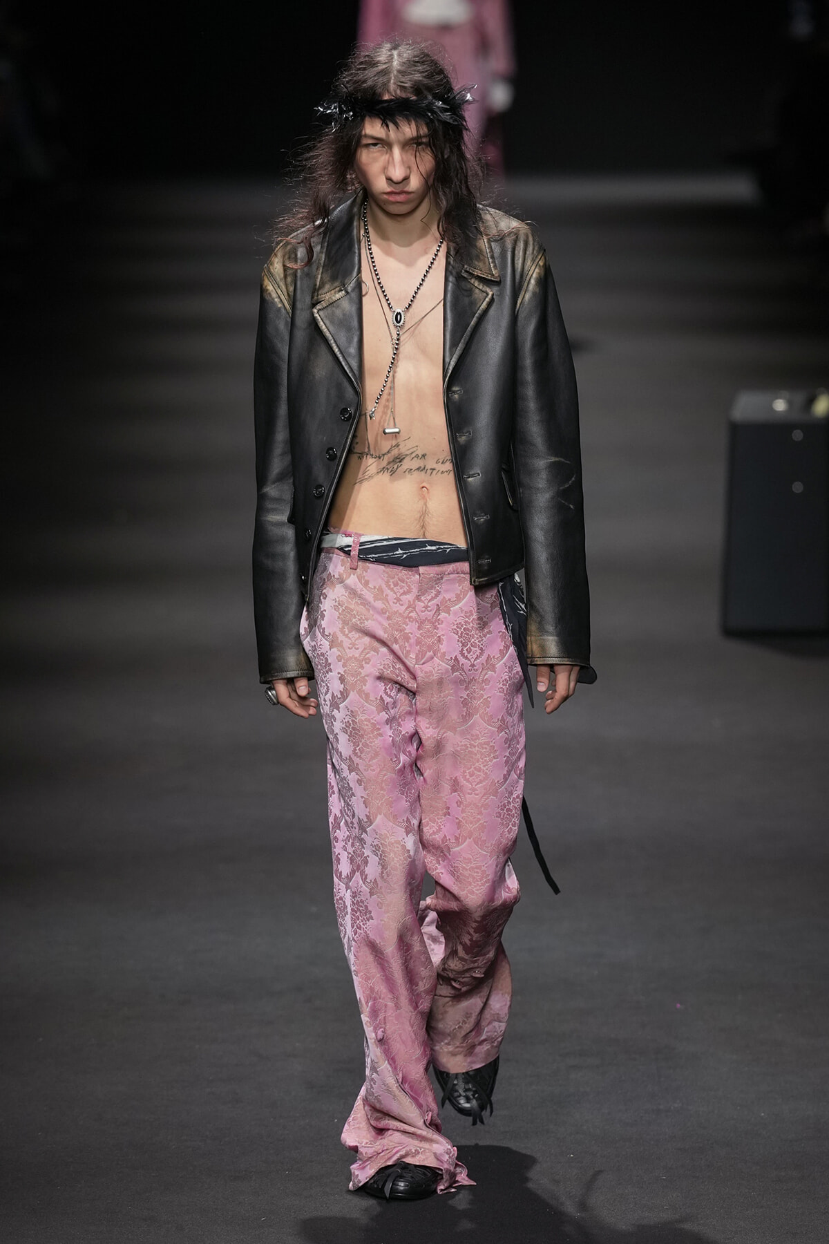 Runway model in a black leather jacket over an open chest, pink damask-patterned pants, and layered necklaces walking down the runway.