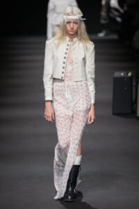 Model walks a runway in a white cropped military-style jacket over a white floral jumpsuit with pink flowers and black boots.