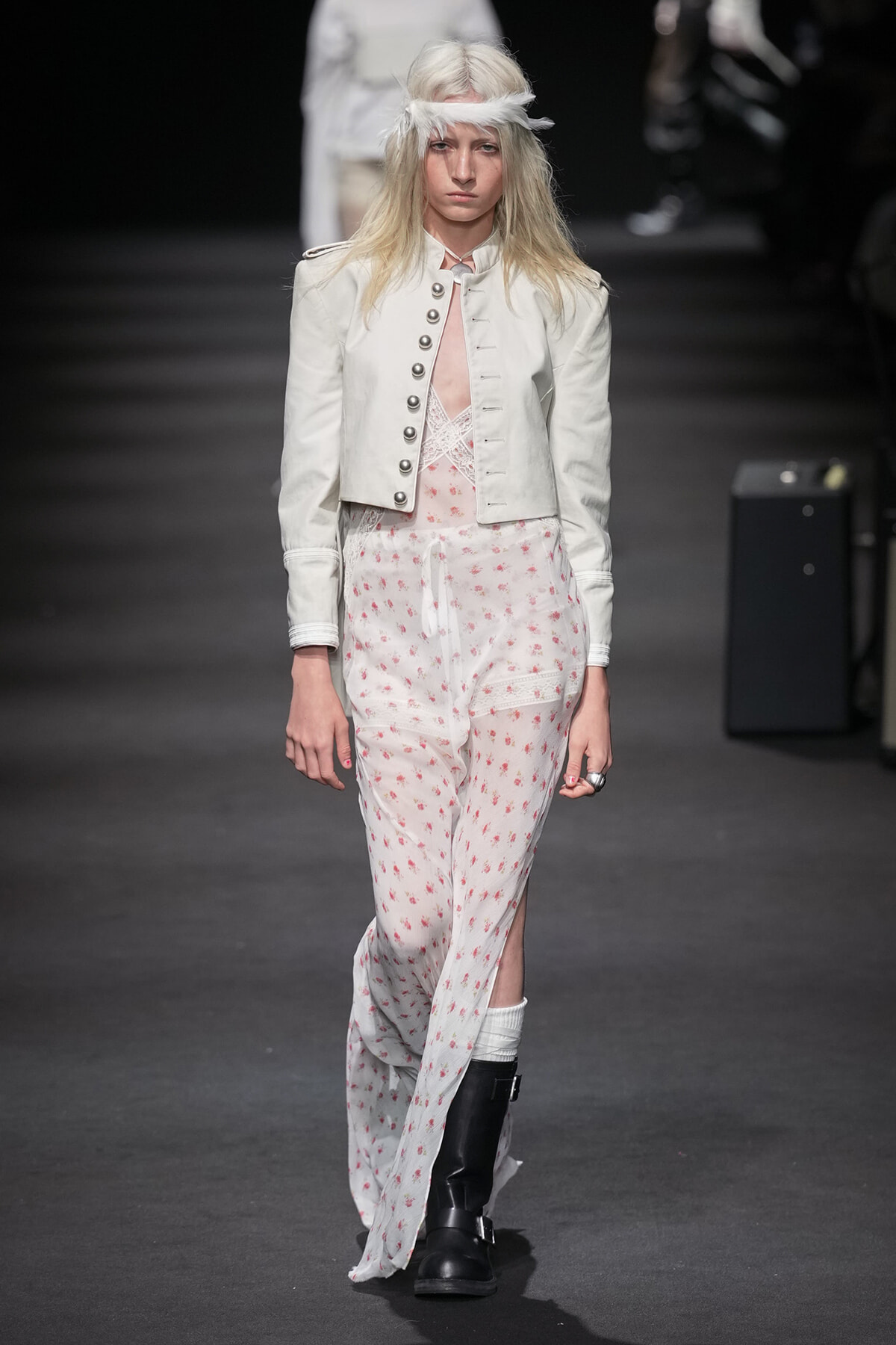 Model walks a runway in a white cropped military-style jacket over a white floral jumpsuit with pink flowers and black boots.