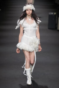 Model walking a runway in an all-white feathered outfit with a fluffy headband and white lace-up boots.