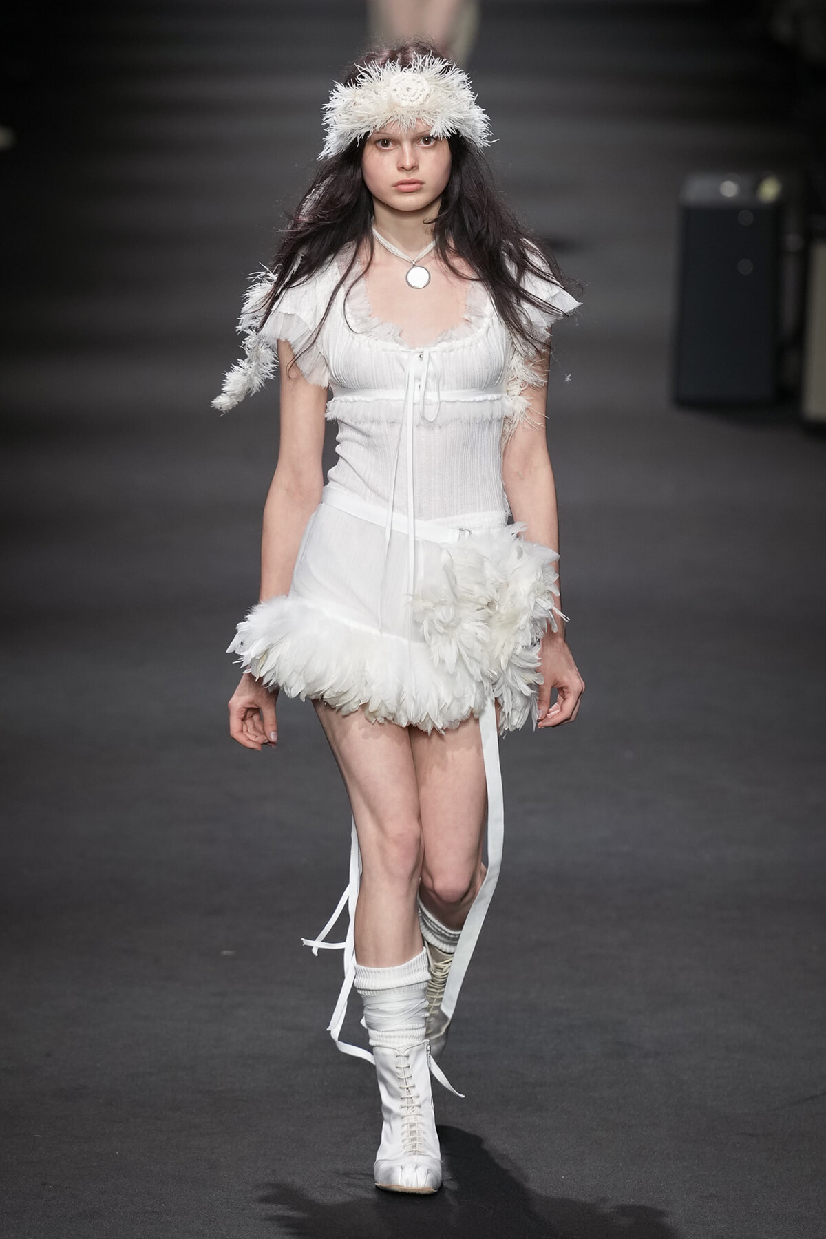 Model walking a runway in an all-white feathered outfit with a fluffy headband and white lace-up boots.