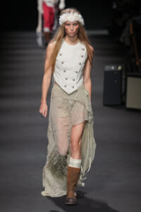 Model on a runway wearing an ivory double-breasted vest with metal buttons over a sheer lace skirt, brown boots, and a white feather headband.