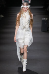 Female model walks the runway in a white, layered dress with a feather scarf and matching feather headband.