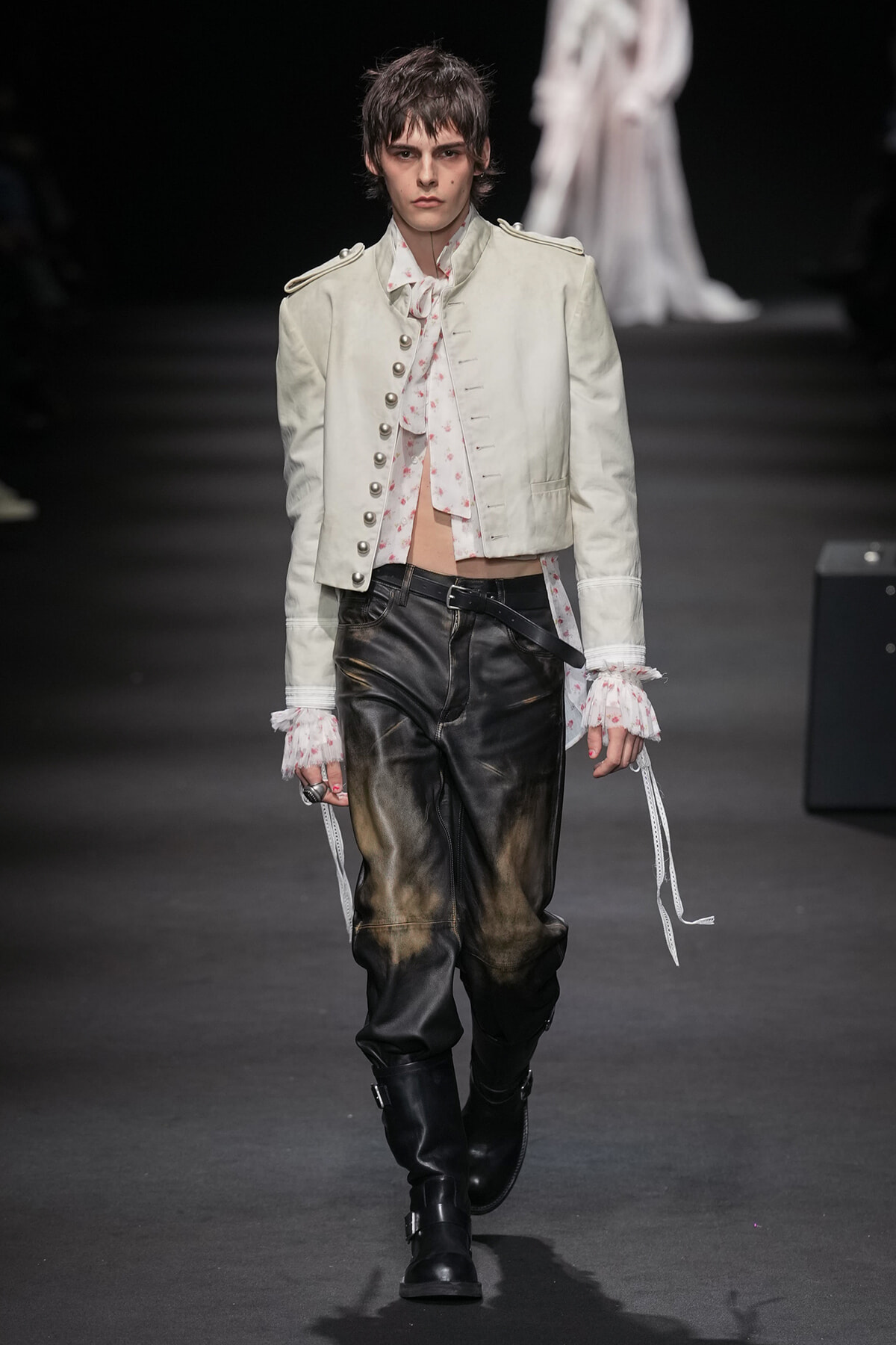 Model walking a runway in a cropped white military-style jacket with floral ruffles, black distressed leather pants, and knee-high boots.