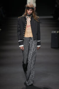 Runway model in a black military-style jacket open to reveal a torso with red 'Dream' text, paired with gray floral wide-leg pants and a feather headband.