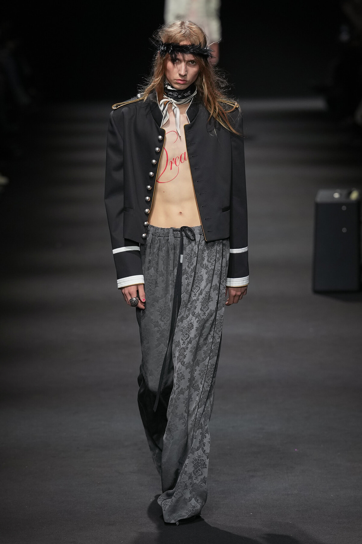 Runway model in a black military-style jacket open to reveal a torso with red 'Dream' text, paired with gray floral wide-leg pants and a feather headband.