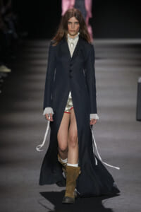 Model walks a runway in a long dark pinstripe coat over a frilled white blouse, metallic shorts, and tan boots.