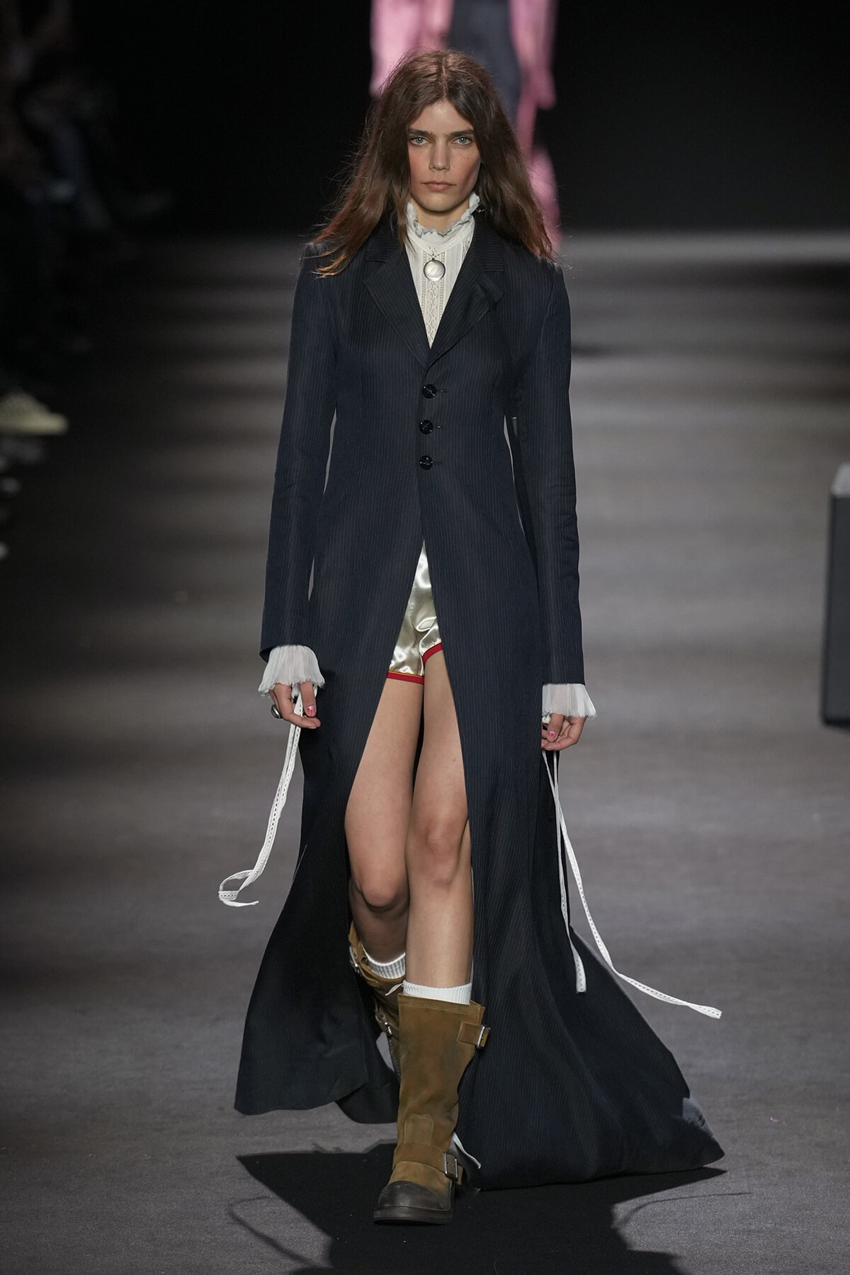 Model walks a runway in a long dark pinstripe coat over a frilled white blouse, metallic shorts, and tan boots.