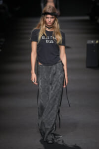 Runway model wearing a black graphic T‑shirt tucked into a long gray damask-pattern skirt with fringe ties, a feathered headband, and a choker.