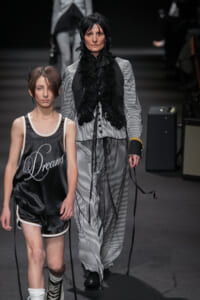 Two models walk a fashion runway: a woman in a black-and-white pinstripe suit with a feathered black scarf, and a young model in a black satin basketball-style outfit that reads 'Dreams'.