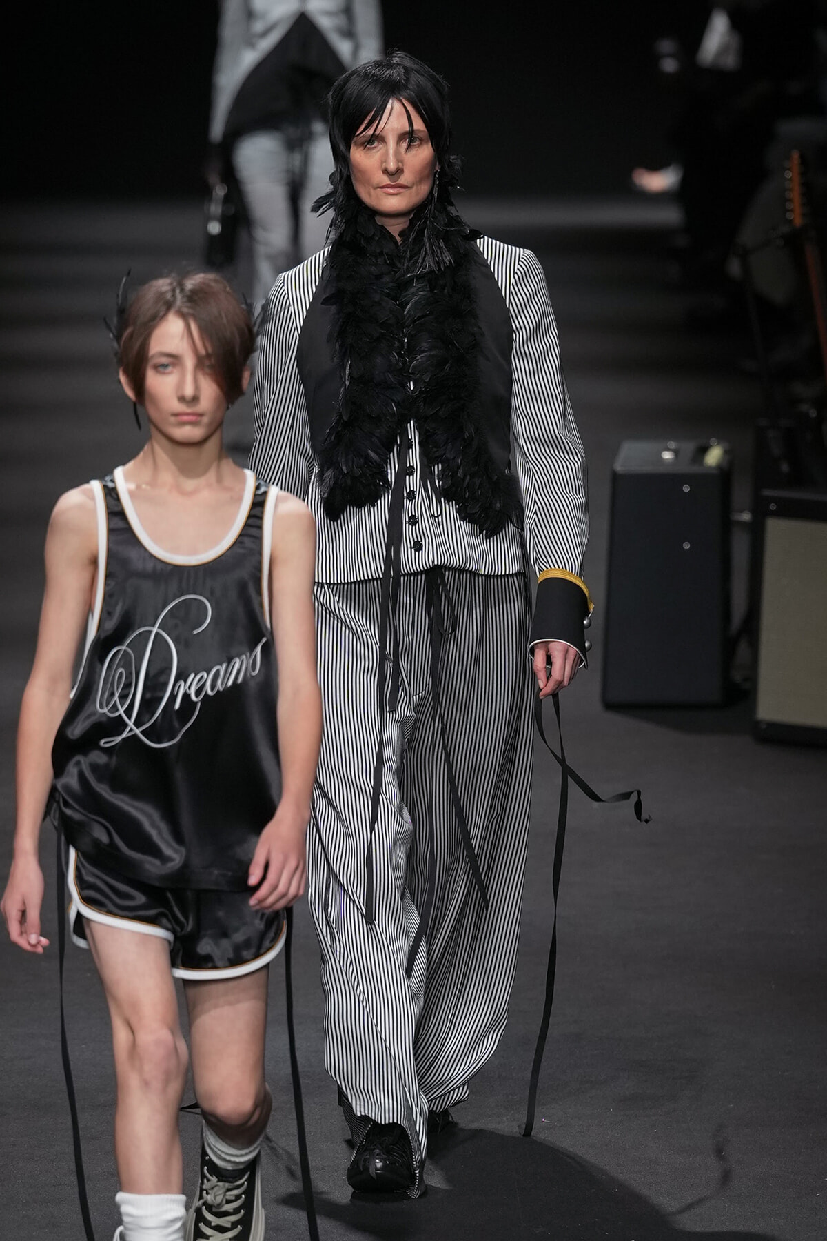 Two models walk a fashion runway: a woman in a black-and-white pinstripe suit with a feathered black scarf, and a young model in a black satin basketball-style outfit that reads 'Dreams'.