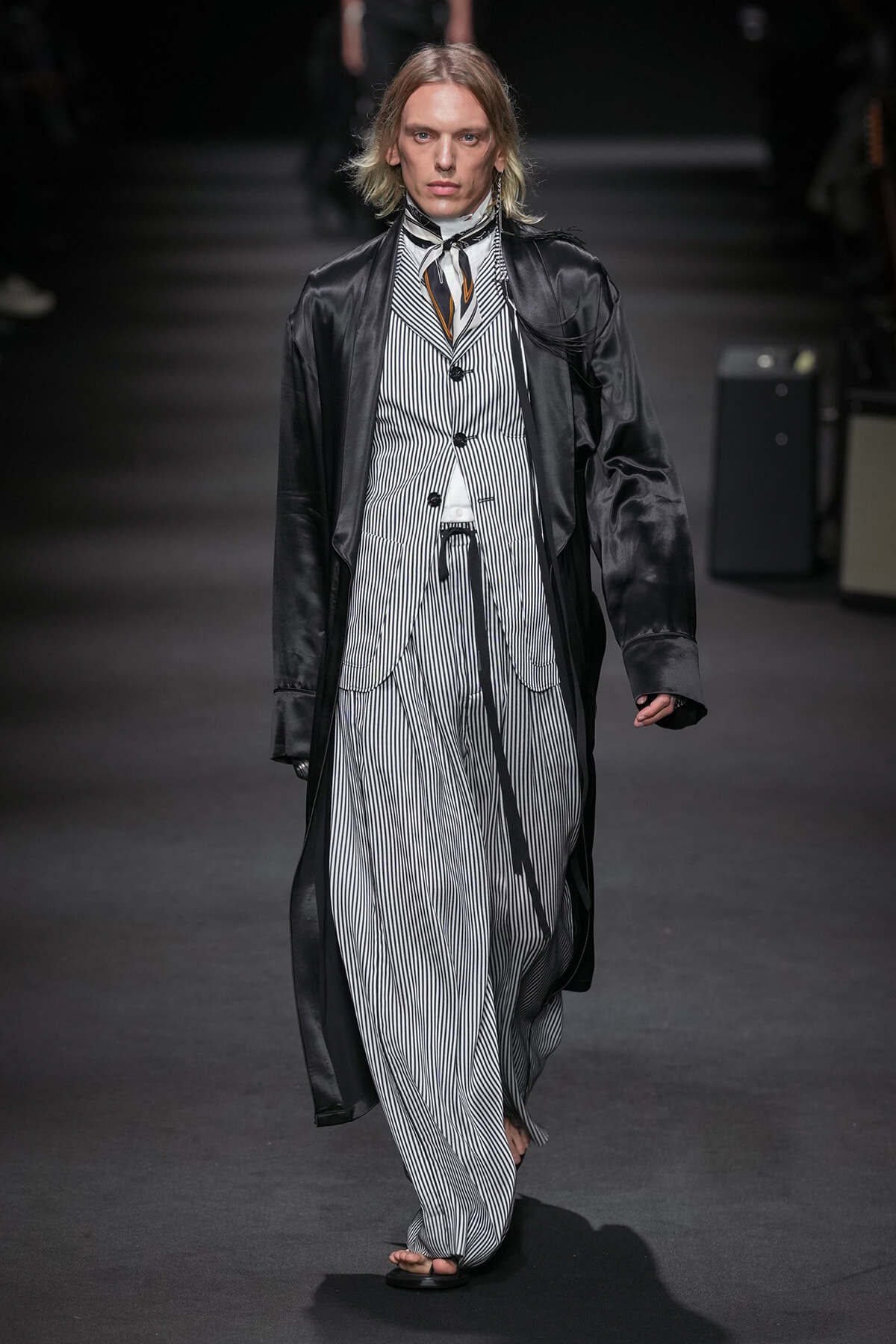 Male model walks a runway in a black satin long coat over a white-and-navy pinstriped suit with a matching waistcoat and scarf or necktie accessory.