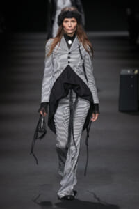 Model walking runway in a black-and-white vertically striped suit with layered draped black fabric and a black headband.
