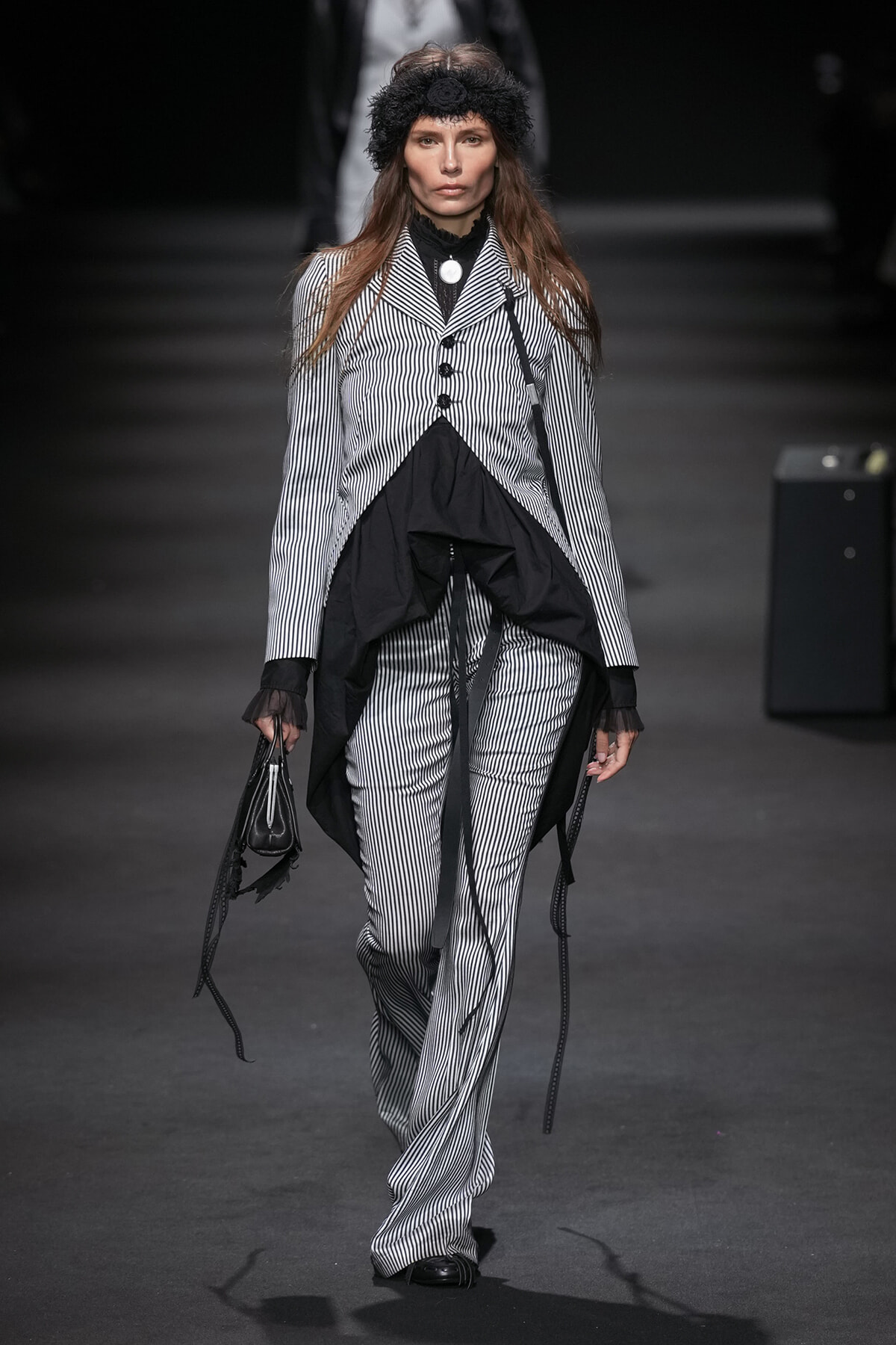 Model walking runway in a black-and-white vertically striped suit with layered draped black fabric and a black headband.