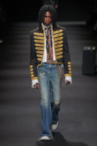 Male model on a runway wearing a black military-style jacket with gold braids, a scarf, distressed blue jeans, a brown belt, and white sneakers.