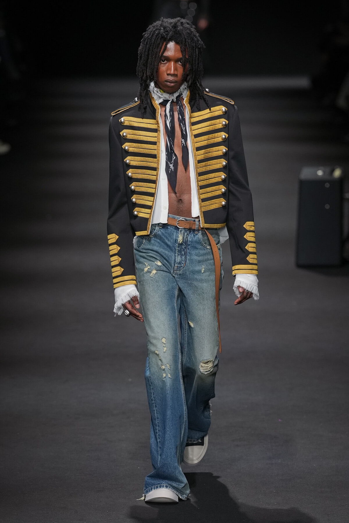 Male model on a runway wearing a black military-style jacket with gold braids, a scarf, distressed blue jeans, a brown belt, and white sneakers.