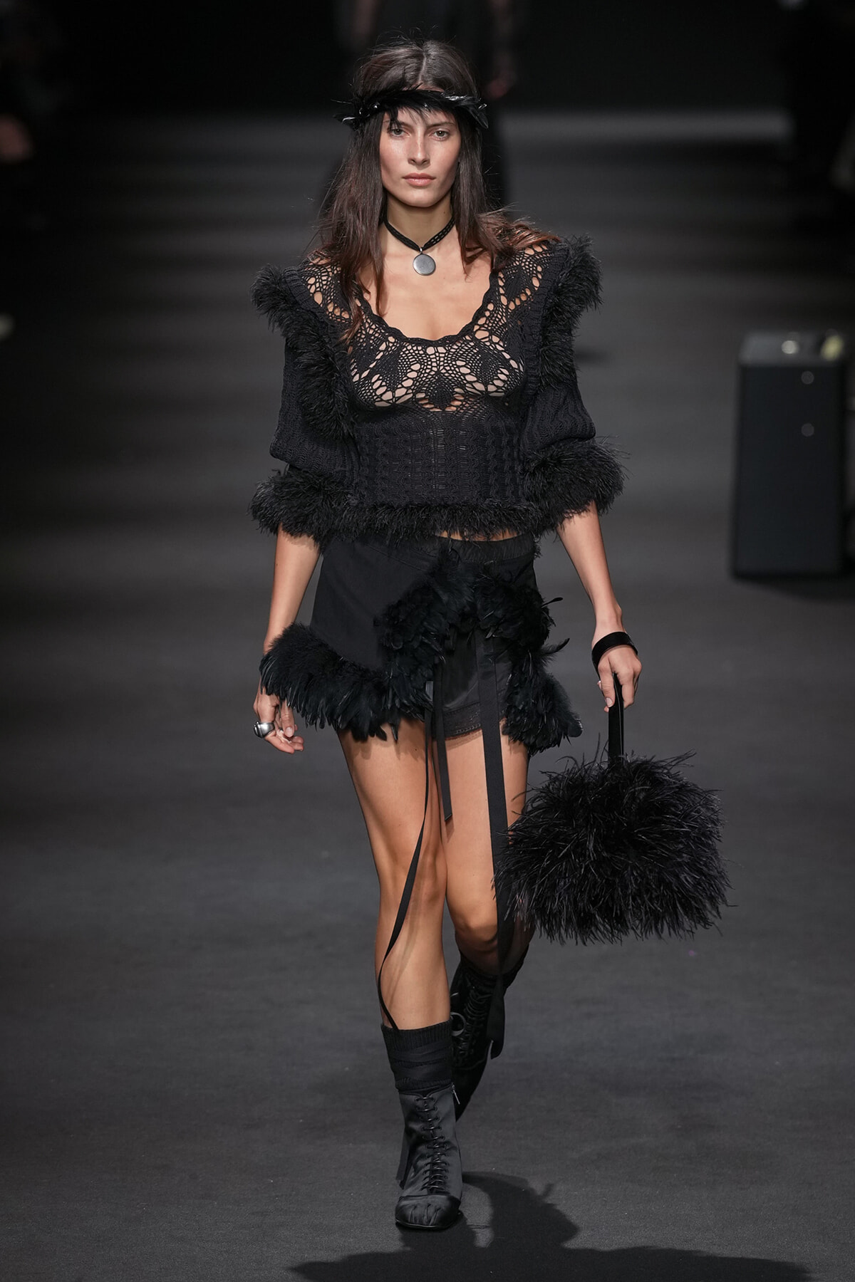 Model walking a runway in a black crochet top with feathered sleeves and hem, paired with black shorts and boots, carrying a fluffy black handbag.