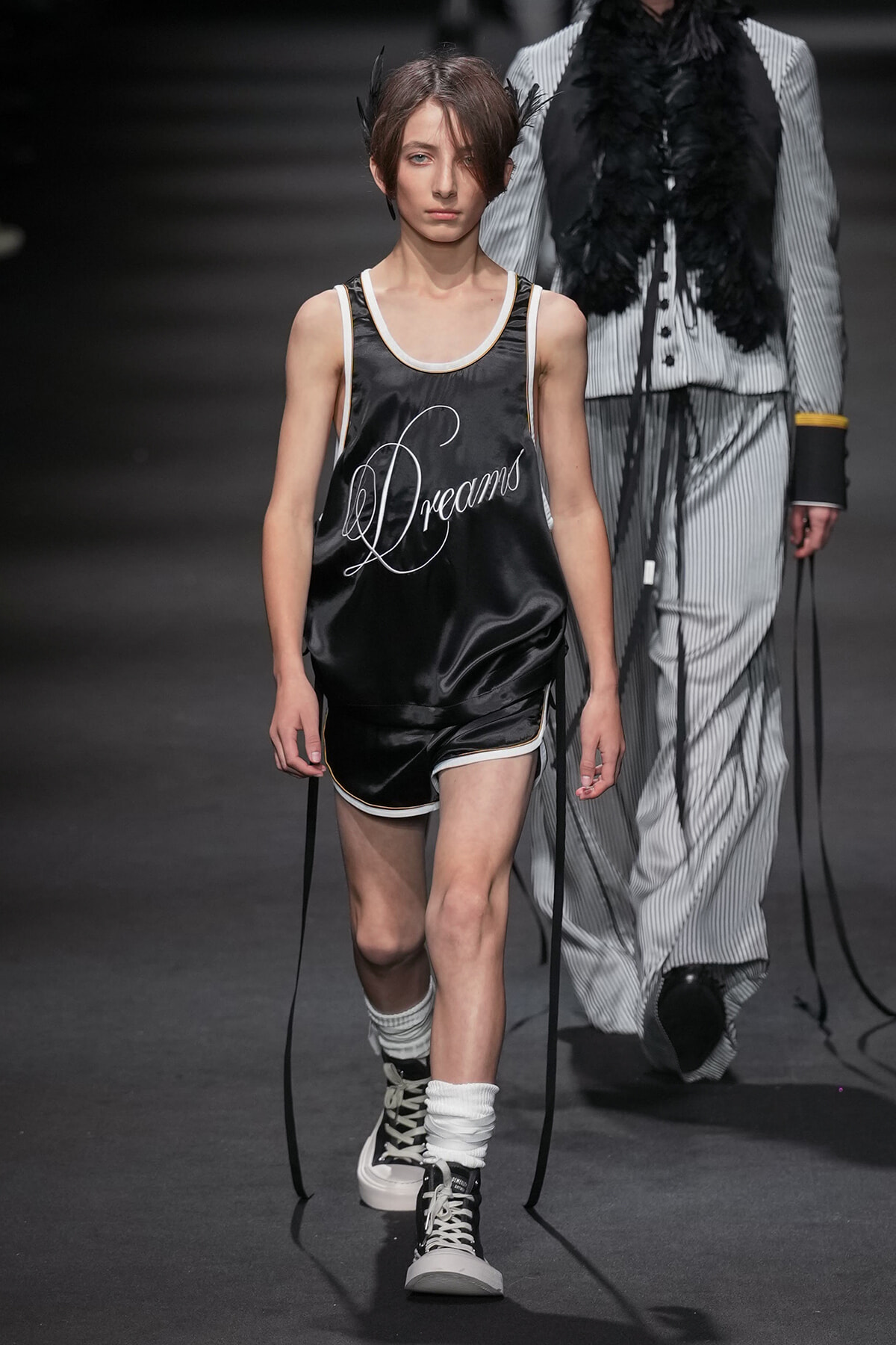 Model walks a fashion runway wearing a black satin basketball-inspired set with white trim and the word 'Dreams' scripted on the jersey, paired with black-and-white high-top sneakers and white leg warmers.