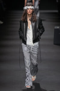 Runway model wearing a black leather jacket over a white blouse and vertically striped pants, with a white feather headband.