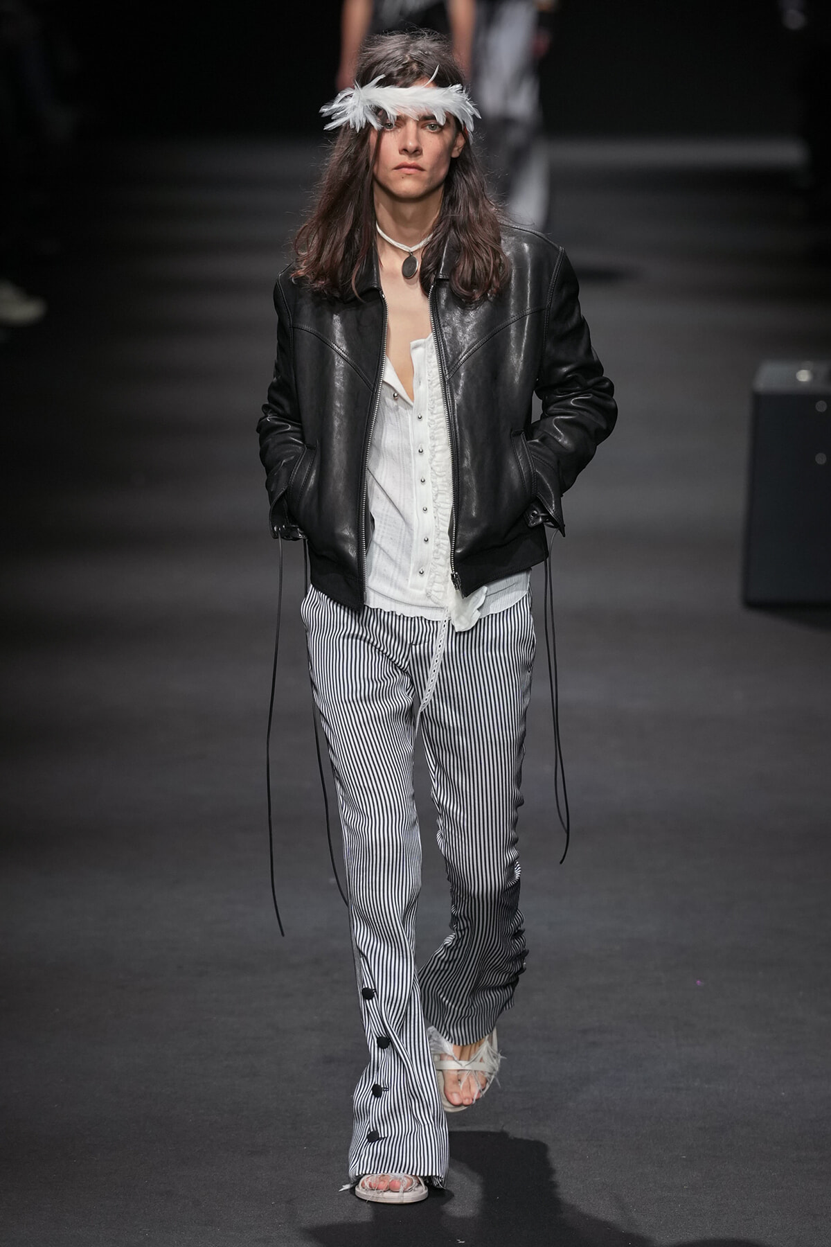 Runway model wearing a black leather jacket over a white blouse and vertically striped pants, with a white feather headband.