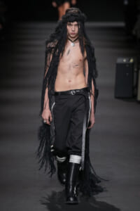 Male model walking runway topless in a black fringe cape, black pants with white stripes, and tall boots; he wears a circular pendant necklace.