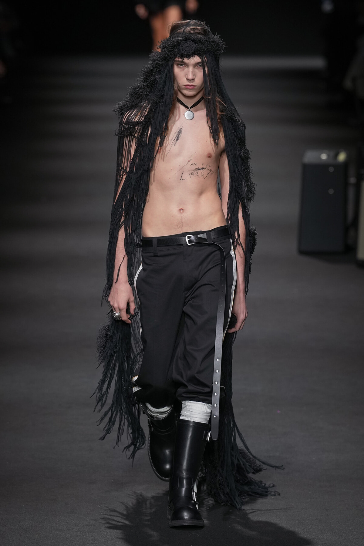 Male model walking runway topless in a black fringe cape, black pants with white stripes, and tall boots; he wears a circular pendant necklace.