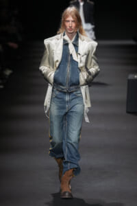 Female model on a fashion runway wearing a metallic silver oversized blazer over a denim jacket and jeans, with a white neck scarf and brown boots.